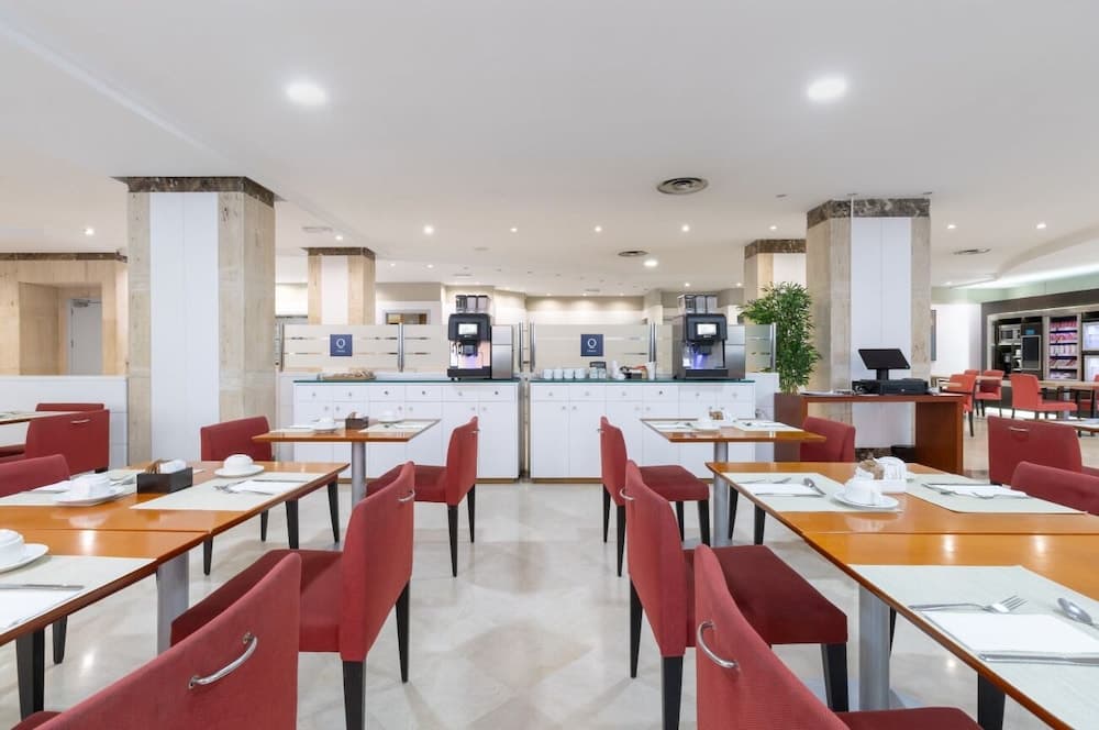 A bright, modern hotel dining area features neatly arranged tables with red chairs, coffee machines, and a welcoming atmosphere ideal for travelers seeking a comfortable breakfast spot.