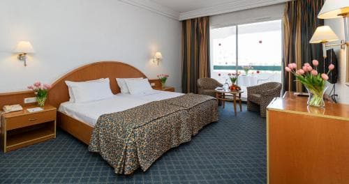A cozy hotel room features a neatly made bed with leopard print bedding, a balcony view through large windows, and vibrant floral arrangements, creating a welcoming atmosphere ideal for travelers seeking comfort and relaxation.