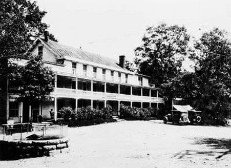 A serene black-and-white scene captures a historic two-story inn surrounded by lush trees, featuring a classic car parked nearby, ideal for travelers exploring vintage architecture and rural charm.