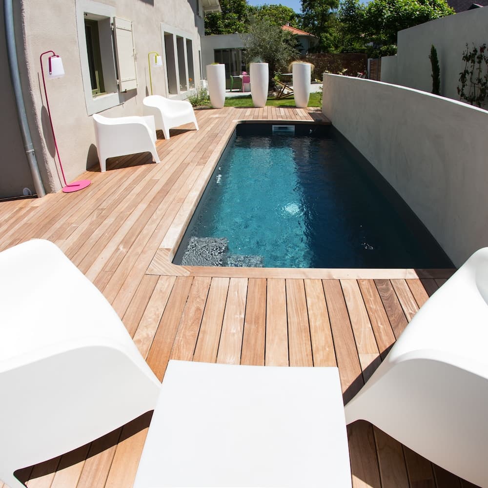 A serene backyard oasis features a sleek rectangular pool surrounded by a wooden deck, white modern furniture, and minimalist decor, perfect for a relaxing vacation retreat.