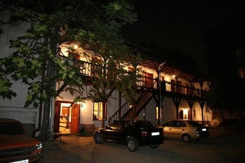 A cozy, warmly lit traditional building with wooden balconies stands invitingly at night, surrounded by parked cars under the shade of trees, ideal for travelers seeking a charming accommodation experience.
