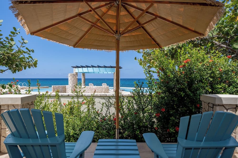 A serene coastal patio features blue Adirondack chairs under a straw umbrella, surrounded by lush greenery and overlooking a tranquil ocean vista, ideal for a relaxing tropical getaway.