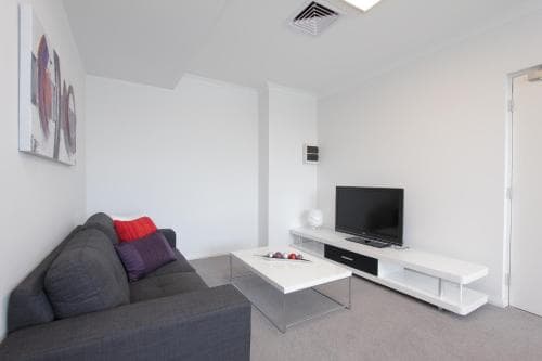 A modern, minimalist living room with white walls, a sleek sofa adorned with colorful cushions, a flat-screen TV on a contemporary stand, and abstract wall art creates a cozy yet stylish atmosphere ideal for short-term rentals or vacation stays.