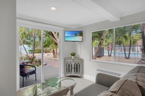 A cozy living room with large windows overlooking a serene beachfront patio, featuring comfortable seating, a TV displaying tropical scenery, and lush greenery outside, ideal for a relaxing vacation retreat.