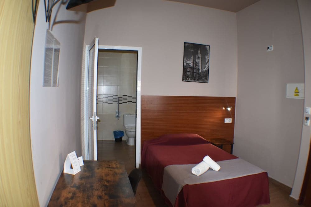 A cozy hotel room features a neatly made bed with red linens, a small wooden desk, an open door to a tiled bathroom, and a framed architectural print on the wall, ideal for travelers seeking simplicity and comfort.