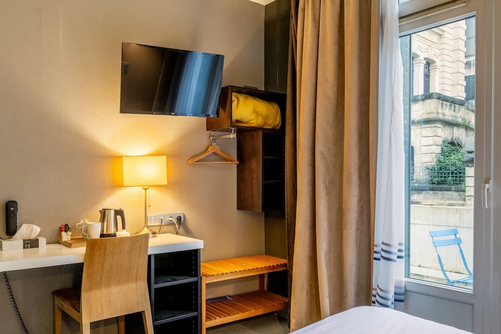 A cozy hotel room features a warm-lit desk area with a kettle and lamp, a mounted TV above a wooden storage unit, and a window view of historic architecture, ideal for travelers seeking comfort and convenience.