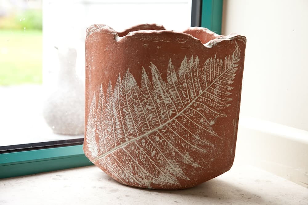 A rustic terracotta vase adorned with white fern imprints sits on a windowsill, bathed in soft natural light, creating a serene indoor decor scene ideal for home aesthetic inspiration.