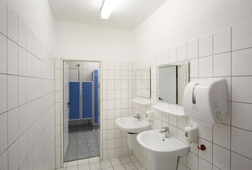 A clean, well-lit public restroom with white tiled walls, blue stalls, two sinks, mirrors, and a paper towel dispenser, ideal for travelers seeking basic facilities during their journey.