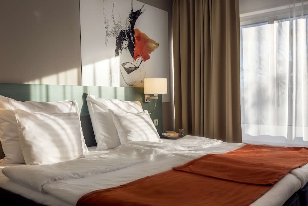 A cozy hotel room bathed in soft sunlight features a neatly made bed with white linens and an orange throw, framed by modern art on the wall and a large window with sheer curtains, ideal for travelers seeking comfort and style.