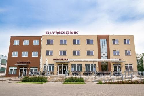 A modern hotel named "OLYMPIONIK" with a welcoming facade, outdoor seating area, and clear blue skies creates an inviting atmosphere ideal for travelers seeking comfortable accommodations and dining options.