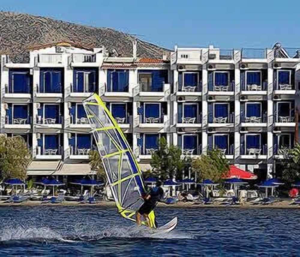 A windsurfer glides across the water in front of a vibrant beachside hotel with blue shutters, surrounded by lounge chairs and umbrellas, ideal for adventure seekers looking for coastal activities.