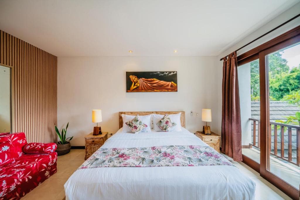 A serene tropical bedroom features a cozy bed adorned with floral bedding, a vibrant red sofa, and a large window opening to lush greenery, ideal for travelers seeking relaxation in a resort setting.