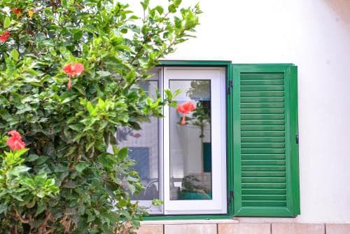 A serene Mediterranean-style window framed in vibrant green shutters is nestled beside lush greenery adorned with red hibiscus flowers, ideal for travelers seeking picturesque coastal charm.