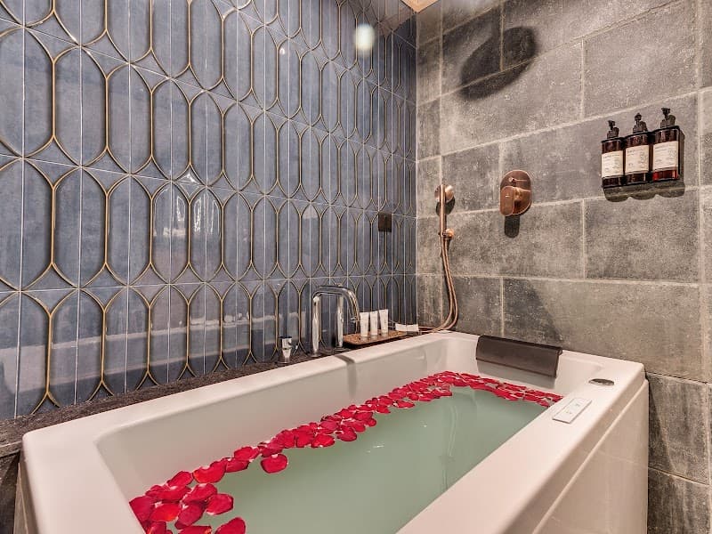 A serene spa bathroom at The Vale Lodge features a luxurious bathtub adorned with red rose petals, elegant blue patterned tiles, and copper fixtures, ideal for travelers seeking relaxation and indulgence.