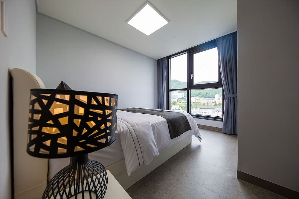 A serene hotel bedroom features a cozy bed by a large window with city views, complemented by a stylish geometric lamp, ideal for travelers seeking comfort and modern design.