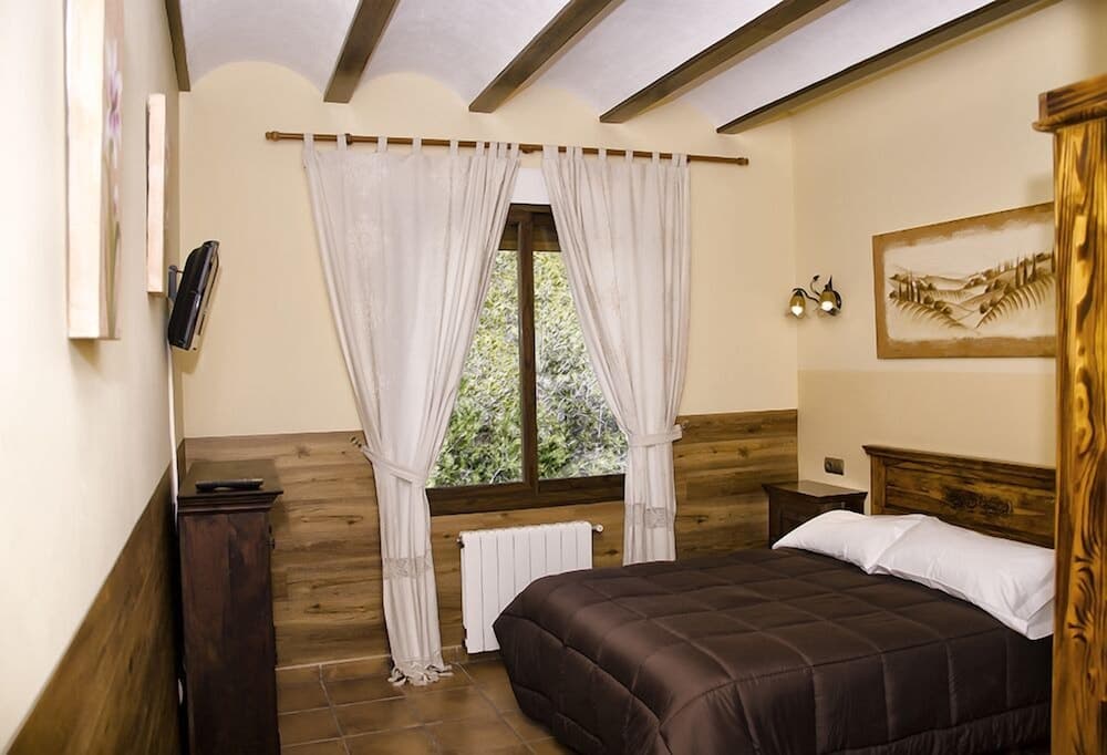 A cozy bedroom with wooden beams, tiled floors, and a window view of greenery offers a serene retreat for travelers seeking relaxation in a rustic setting.