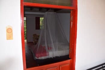 A cozy bedroom with a mosquito net-covered bed framed by a vibrant red window, evoking a serene tropical retreat ideal for travelers seeking relaxation and protection from insects.