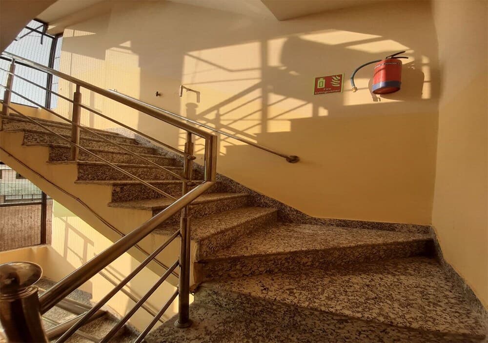 A sunlit indoor staircase with metal railings and a mounted fire extinguisher creates a functional yet serene atmosphere, ideal for exploring urban architecture and safety features during city tours.