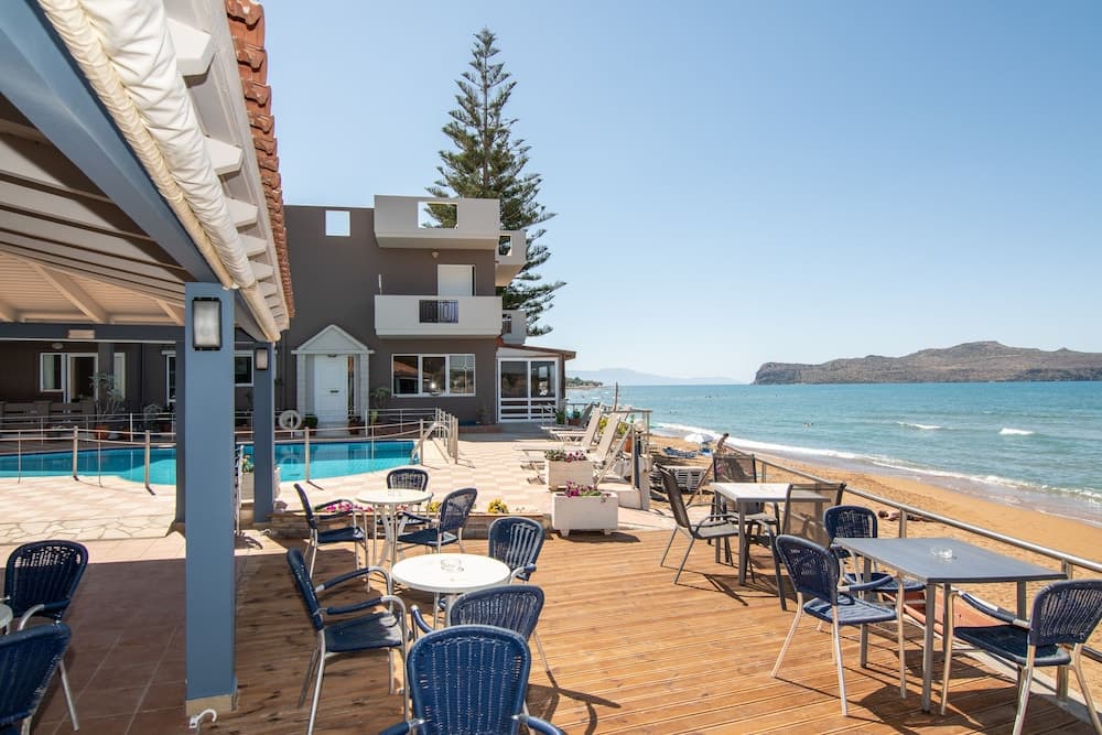A serene seaside café with blue chairs and tables overlooks a tranquil beach and turquoise waters, perfect for travelers seeking relaxation and coastal views.