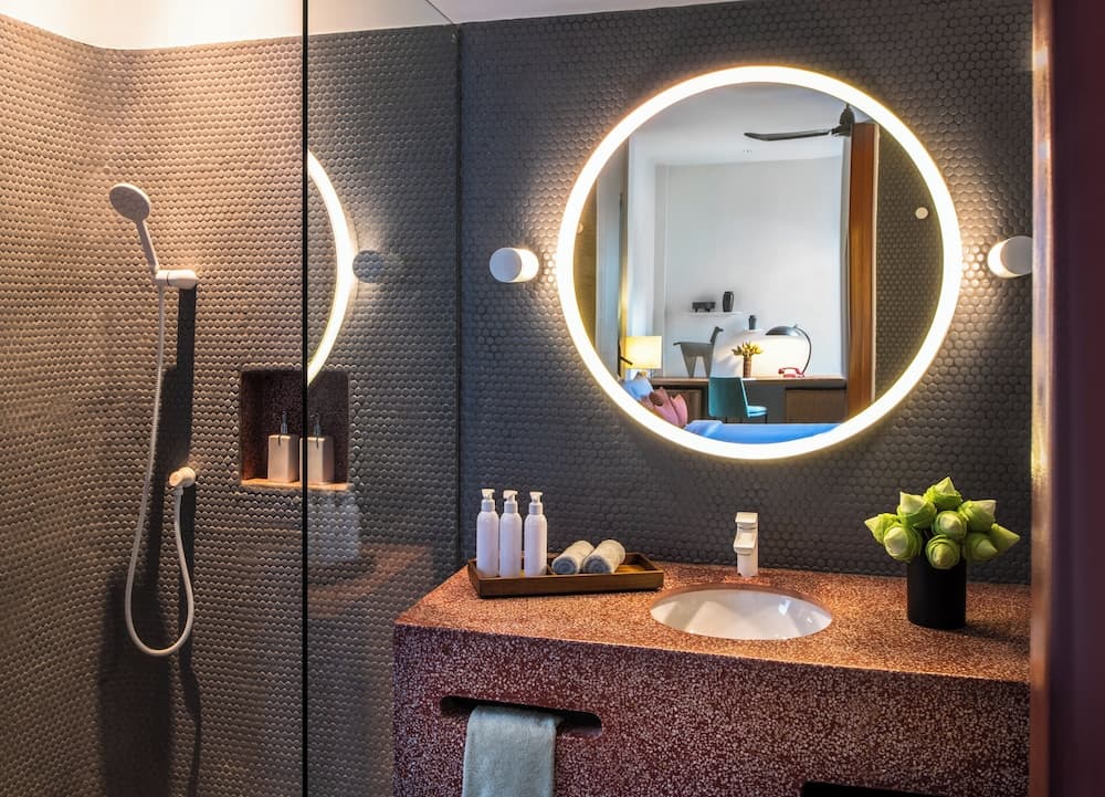 A modern bathroom features a sleek glass shower, a round illuminated mirror reflecting a cozy bedroom, and a stylish vanity with toiletries and greenery, ideal for luxury hotel stays or upscale home designs.