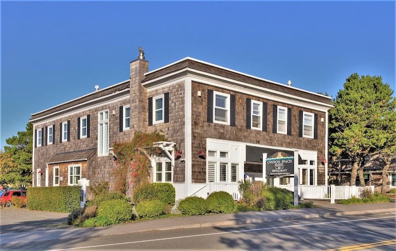 A charming two-story hotel with shingled architecture and lush greenery invites travelers to Cannon Beach, Oregon, offering a cozy retreat under clear blue skies.