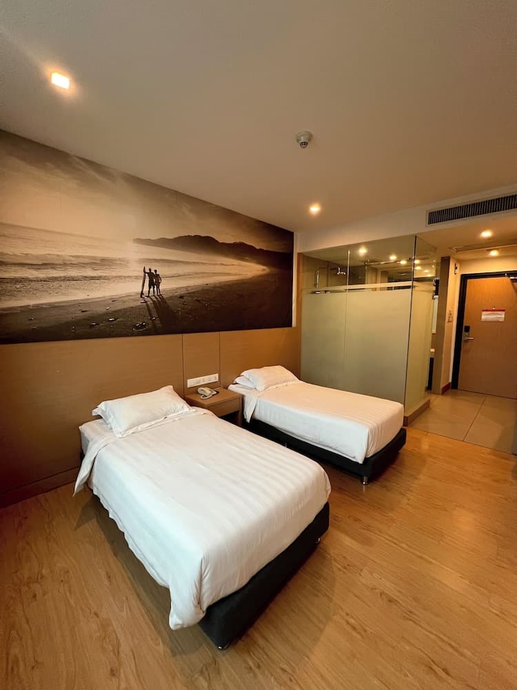 A cozy hotel room features two neatly made beds under a large beach mural, a glass-enclosed shower, and warm wooden flooring, ideal for travelers seeking comfort and relaxation.