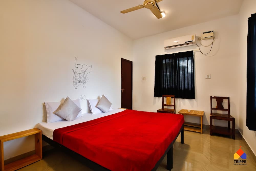A cozy hotel room features a bed with a vibrant red blanket, wooden furniture, black curtains, an air conditioner, and a playful wall drawing, ideal for travelers seeking comfort and simplicity.