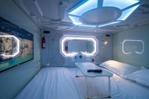 A futuristic capsule hotel room with sleek white decor, illuminated windows, and a cozy bed setup offers a unique and modern accommodation experience for travelers seeking innovative lodging options.
