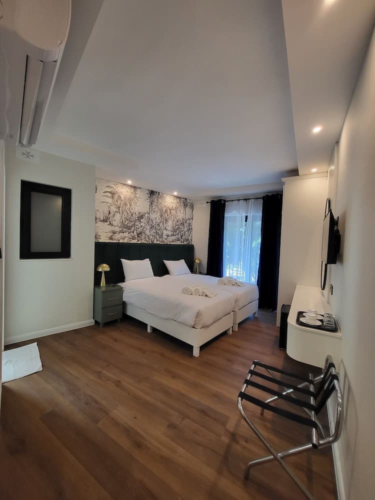 A cozy hotel room features a neatly made bed with white linens, a tropical mural backdrop, wooden flooring, and a small dining area, ideal for travelers seeking comfort and style.