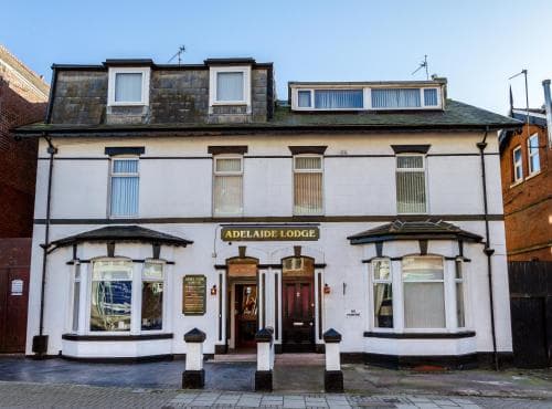 A charming white-painted Adelaide Lodge hotel stands invitingly on a quiet street, featuring classic architecture with bay windows and a welcoming entrance, ideal for travelers seeking cozy accommodations in a quaint urban setting.