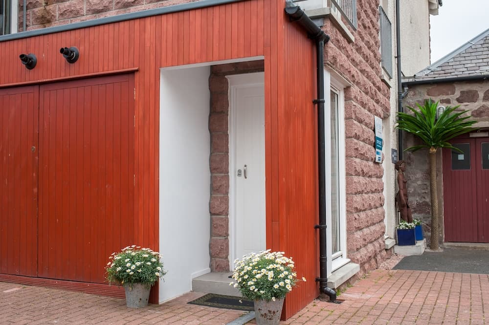 A charming red-painted wooden entrance with white door framed by stone walls, flanked by potted daisies, exudes a cozy village vibe ideal for travelers seeking quaint accommodations.