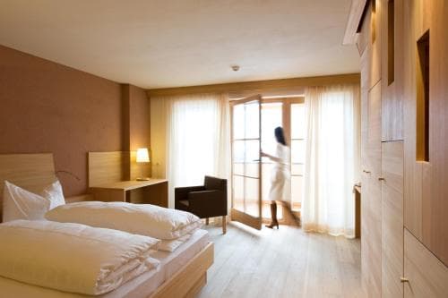A serene hotel room with a neatly made bed, wooden furnishings, and a person stepping onto a balcony through glass doors, ideal for travelers seeking relaxation and comfort.