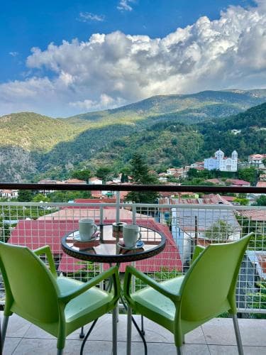 A serene balcony view overlooking lush green hills and quaint rooftops under a partly cloudy sky, featuring green chairs and a coffee table, perfect for a relaxing getaway or vacation rental promotion.