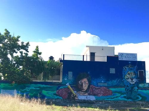 A vibrant mural featuring a girl and a jaguar adorns the wall of Hotel Balché under a clear blue sky, creating an artistic and serene atmosphere ideal for travelers seeking cultural exploration and relaxation.