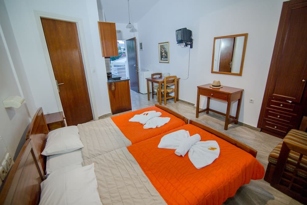 A cozy studio apartment features a neatly arranged bed with orange bedding and towel swans, a small dining area, kitchenette, and wooden furnishings, ideal for travelers seeking comfort in a compact space.