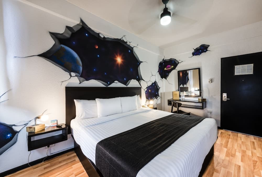 A cozy hotel room features a vibrant cosmic mural breaking through the wall, creating a dreamy atmosphere with a neatly made bed, modern bedside lamps, and a sleek vanity area, ideal for travelers seeking a unique and artistic stay.