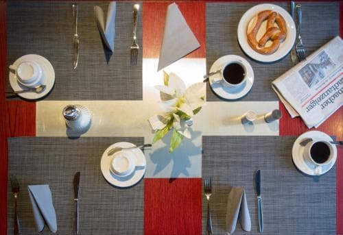 A cozy breakfast table set with coffee cups, a pretzel, newspapers, and elegant white flowers on a patterned placemat, ideal for travelers seeking a charming morning experience in a boutique hotel.