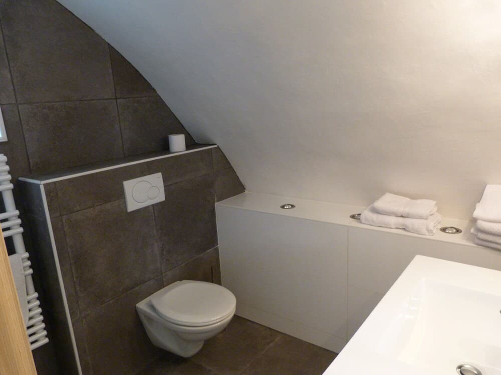A cozy attic bathroom with a slanted ceiling features a modern toilet, sleek white bathtub with neatly folded towels, and dark tiled walls, ideal for travelers seeking compact yet stylish accommodations.
