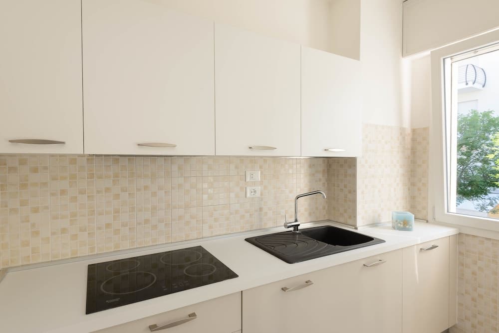 A bright, modern kitchen with sleek white cabinets, mosaic tile backsplash, and natural light from a window offers an inviting space for culinary enthusiasts seeking a clean and functional cooking environment.