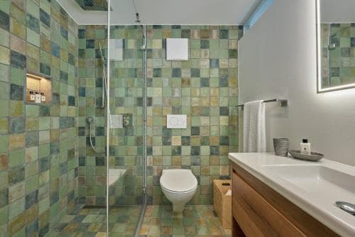 A modern bathroom with vibrant green mosaic tiles exudes a fresh and serene vibe, featuring a sleek shower, toilet, wooden vanity, and neatly arranged toiletries, ideal for travelers seeking comfort and style in their accommodations.