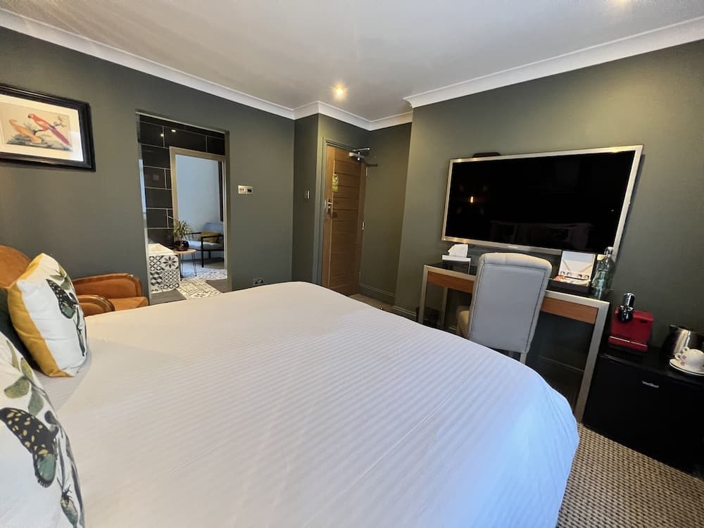 A cozy hotel room with olive green walls, a neatly made bed adorned with floral pillows, a modern desk setup, and a mounted TV creates a serene atmosphere ideal for travelers seeking comfort and relaxation.