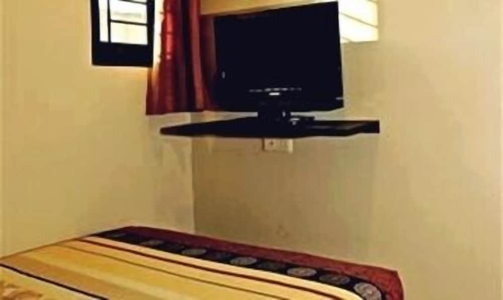 A cozy hotel room corner features a mounted flat-screen TV above a neatly made bed with patterned bedding, set against light walls with a small window and red curtains, ideal for travelers seeking comfort and relaxation.