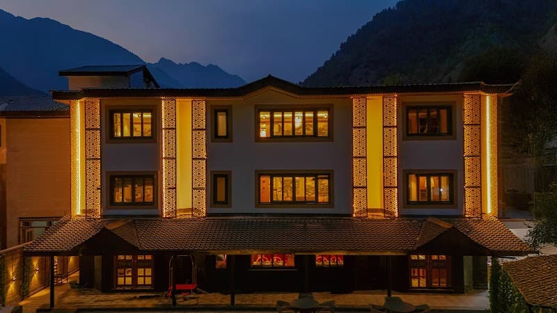 A warmly lit lodge with intricate facade designs stands invitingly against a serene mountain backdrop at dusk, ideal for travelers seeking cozy accommodation in a tranquil valley setting.