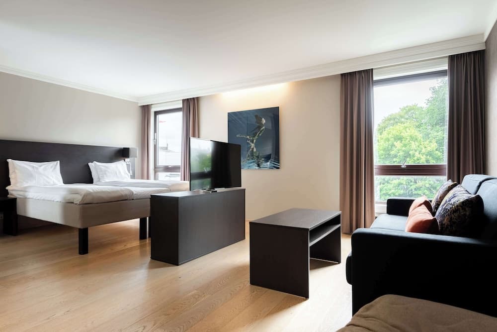 A cozy hotel suite features a neatly made bed, modern furnishings, a flat-screen TV, and large windows offering a serene view of greenery, ideal for travelers seeking comfort and relaxation.