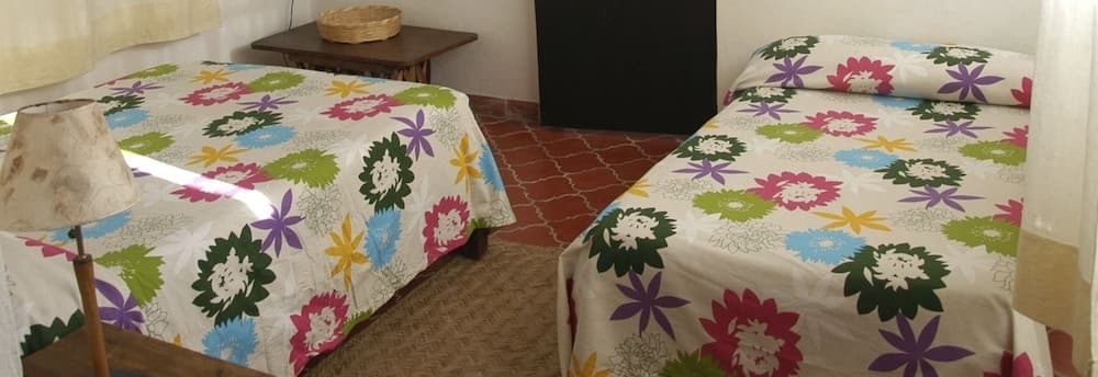 A cozy guesthouse room features two beds adorned with vibrant floral bedding, a rustic lamp, and terracotta flooring, ideal for travelers seeking a charming and comfortable stay.
