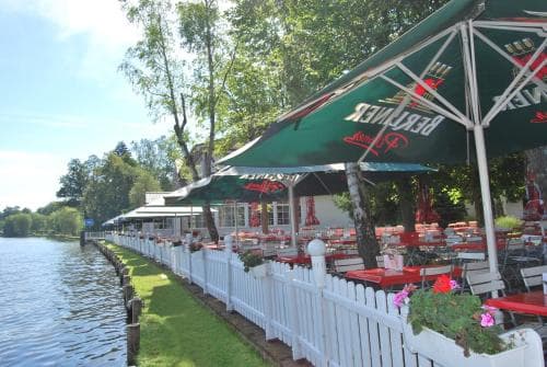 A serene riverside café with vibrant green umbrellas and white picket fences offers a picturesque spot for travelers seeking a tranquil outdoor dining experience amidst lush greenery.