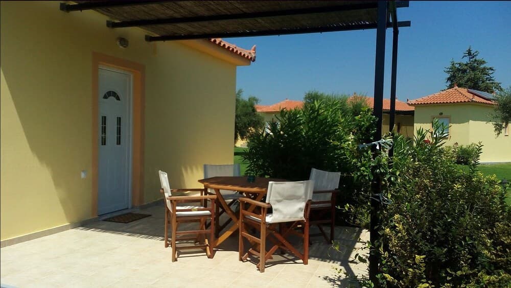 A serene outdoor patio features wooden furniture under a thatched roof, surrounded by lush greenery and vibrant yellow walls, ideal for a relaxing vacation retreat.