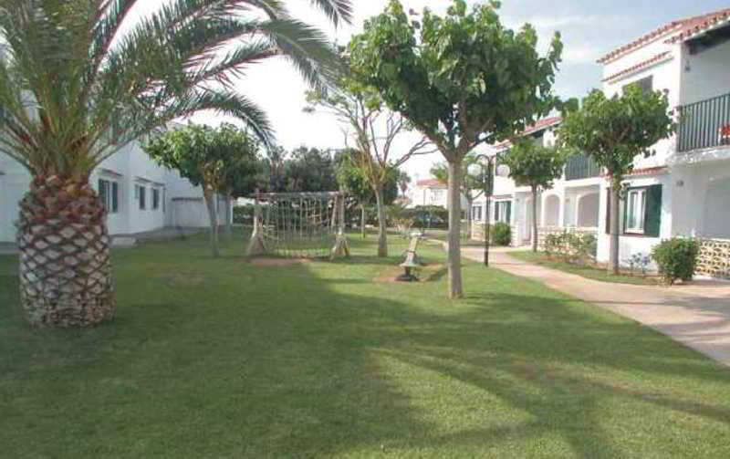 A serene residential area features lush green lawns, palm trees, and a children's playground, ideal for family vacations seeking relaxation and leisure activities.