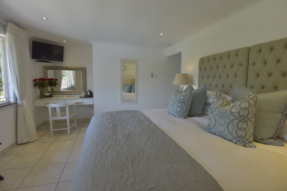 A serene hotel bedroom features a cozy bed with patterned pillows, a stylish vanity with fresh flowers, and soft lighting, ideal for travelers seeking relaxation and comfort.