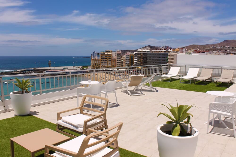 A serene rooftop terrace with lounge chairs, potted plants, and a stunning ocean view offers a relaxing retreat for travelers seeking coastal tranquility amidst urban architecture.